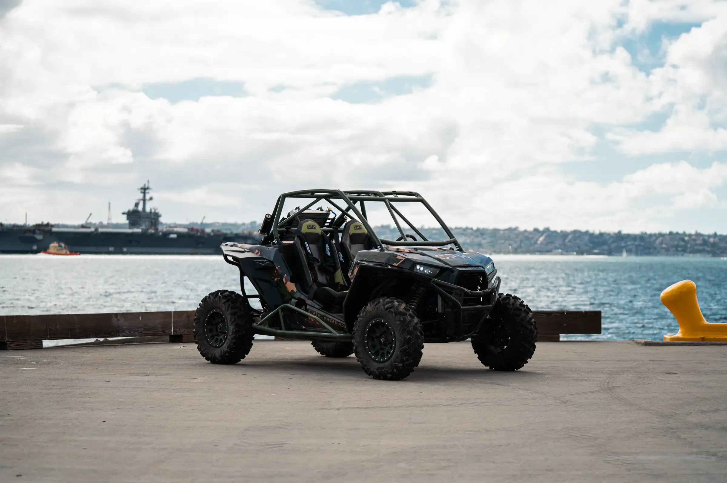 SPM Alpha Prototype electric UTV in olive drab and camouflage on a naval pier, aircraft carrier moored behind it